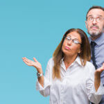 Middle age hispanic couple in love wearing glasses over isolated background clueless and confused expression with arms and hands raised. Doubt concept.